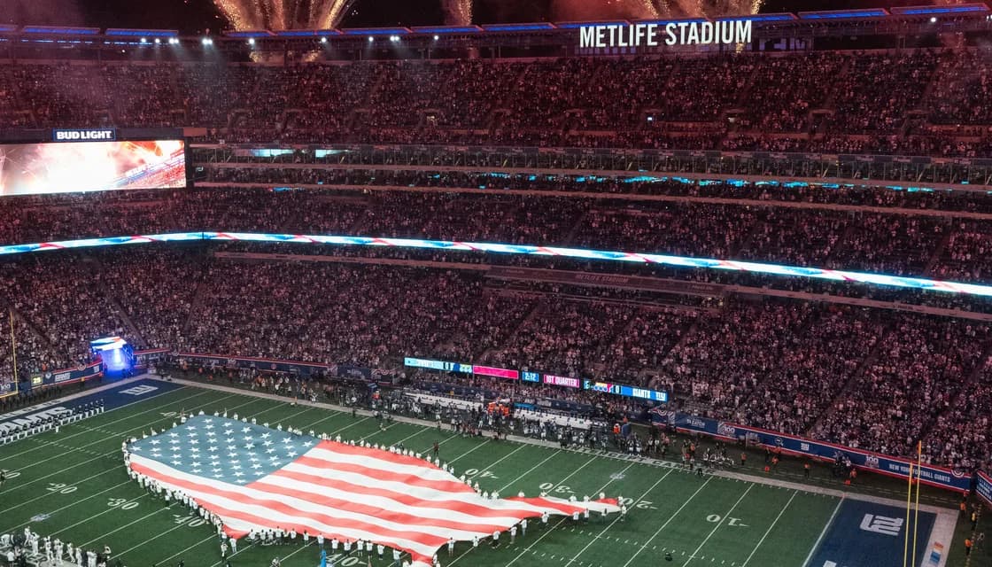 A packed MetLife Stadium at night, a field-sized American flag unfurled across the turf during the pre-game national anthem.