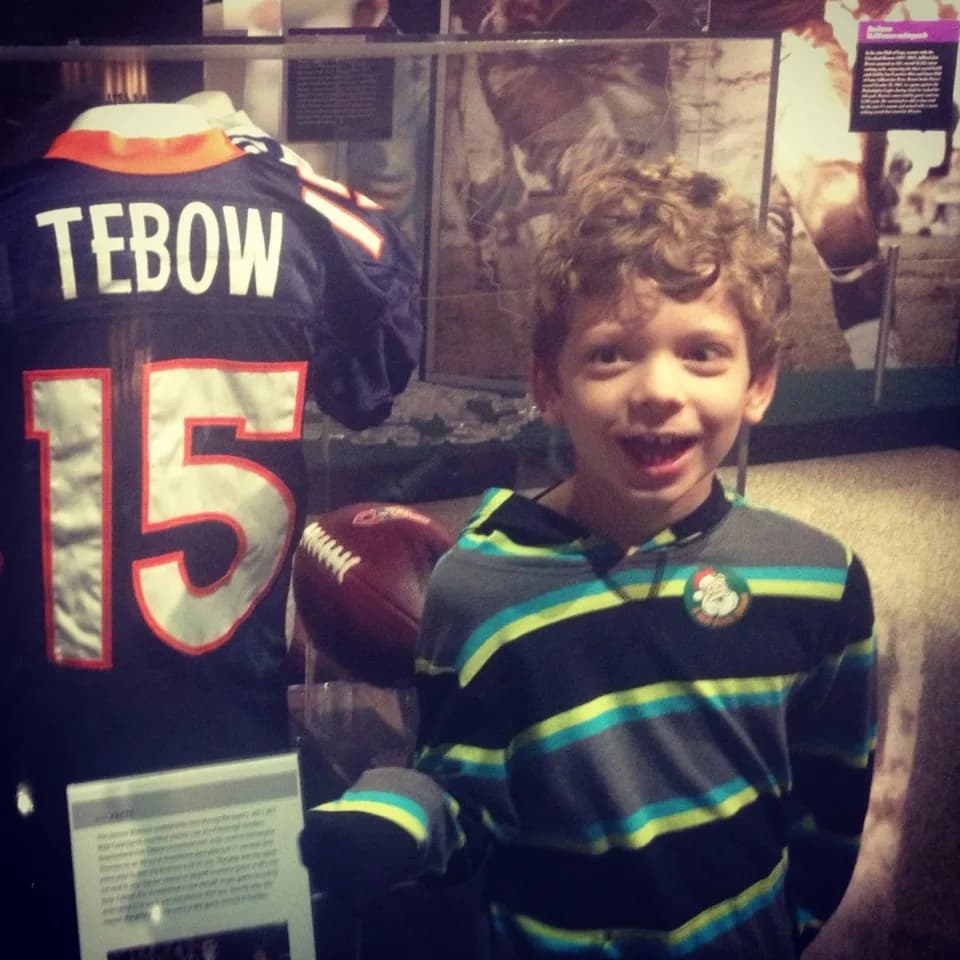 Childhood photo of the author at a football exhibit beside a Tim Tebow jersey.